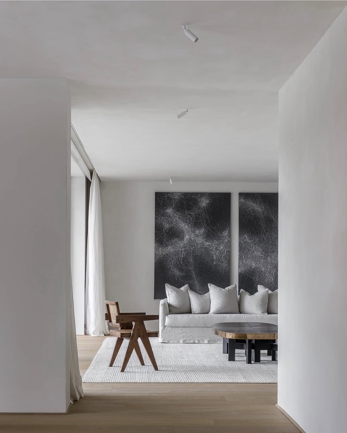 White interior with two large dark abstract paintings above a linen sofa, a cane chair and a round bronze table